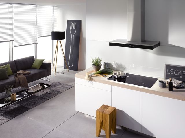view of a modern kitchen and living room area, with the kitchen in the foreground and the living room the background. The kitchen has white cabinets and beige countertops with a black induction cooktop with a stainless steel pan on top. beside the cooktop, is a leek, and a small grocery list chalkboard that says "leeks" on it