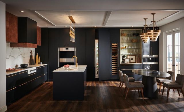 front view of a modern luxury kitchen with a black wall in the back with built in wolf ovens and sub zero refrigerators, and an island with white marble countertops in front