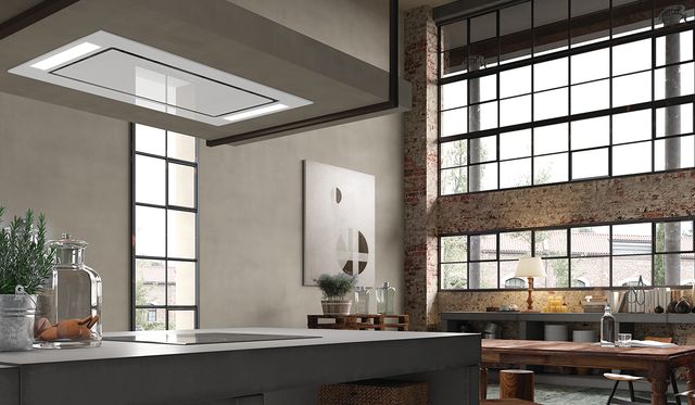 angular view of a kitchen island with a ceiling mounted range in the foreground and an industrial style studio living room in the background