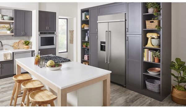 angled view of a kitchen with white wall and grey cabinets. A white kitchen island sits in the middle and a large side by side built in refrigerator sitting behind it in the cabinetry