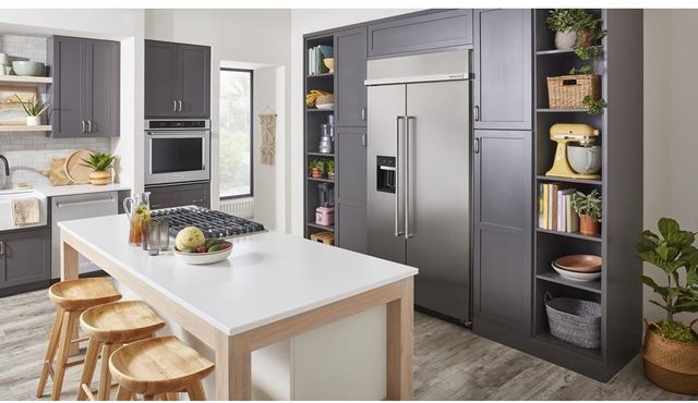 front view of a modern kitchen with a white island, grey cabinetry, and stainless steel appliances. Built into the wall of cabinetry behind the island is a large kitchenaid refrigerator
