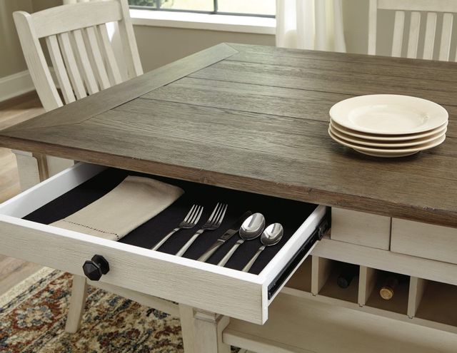 closeup view of a wooden dining table with a drawer, pulled out to show silverware and napkins inside, with wine storage in the table on the right