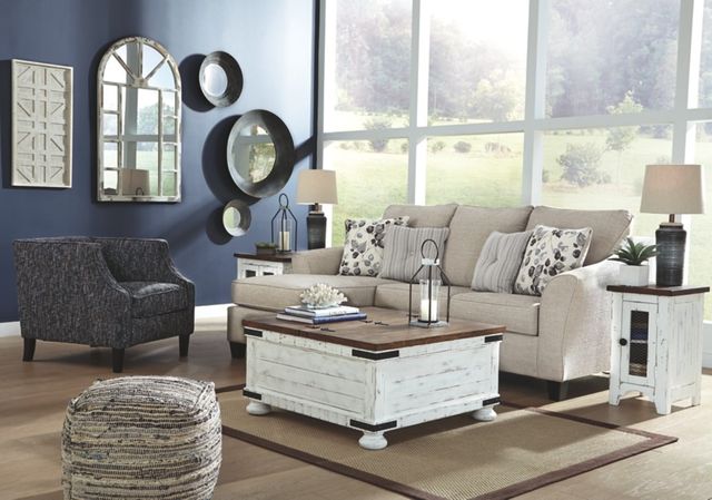 view of a modern living room with the Benchcraft Abney 7-Piece Living Room Set of furniture and decor throughout. Includes a beige sofa, a grey accent chair, white and brown coffee and end tables, a blue wall with various mirrored accents, and a large window wall in the background