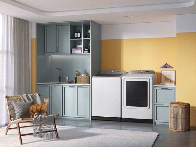 front view of a laundry room with yellow walls and grey cabinets, with a steam washer and dryer centered on the wall. A small seating area peaks on the left, with an orange cat on the chair.