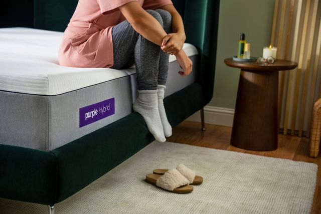 side view of a woman sitting on the edge of a purple mattress on an emerald green bed frame, with her slippers on the beige rug on the floor