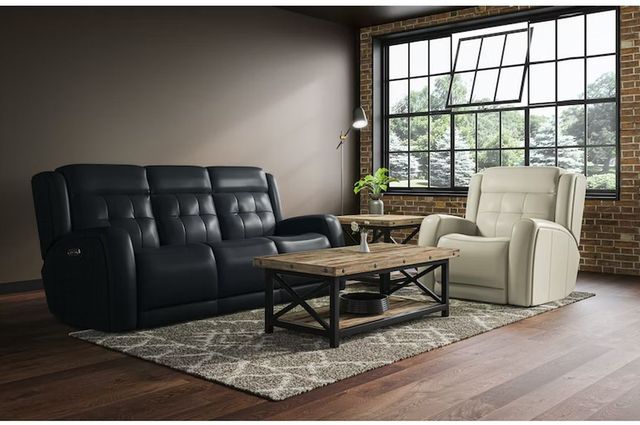 front view of a modern living room with a black sofa and white recliner, with a beige and brick wall with large windows in the background