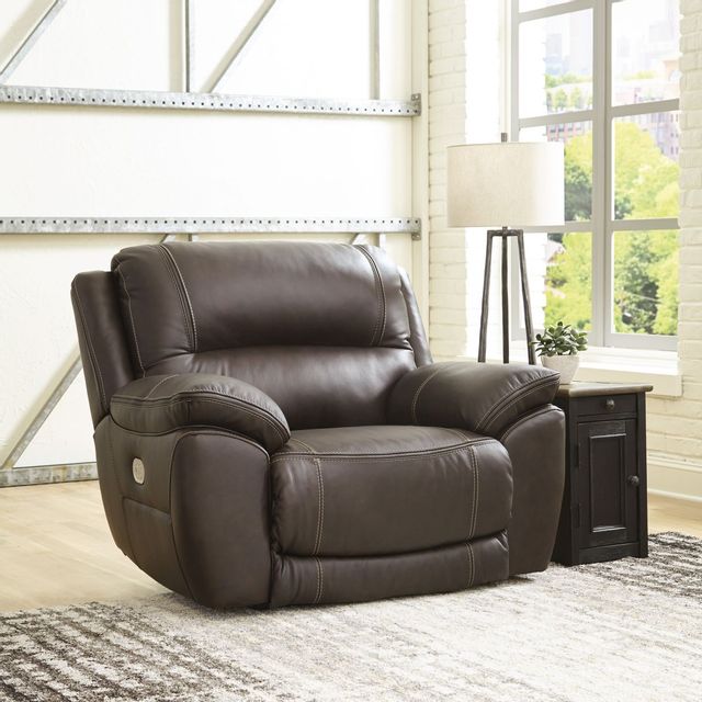 front view of a brown leather oversized recliner in a modern white room corner with a side table, lamp, and window in the background