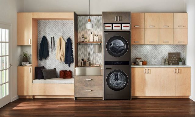 front view of a modern laundry room with an lg washtower and a sink in a built in wall storage feature made of light gray wood on a white tile backsplash