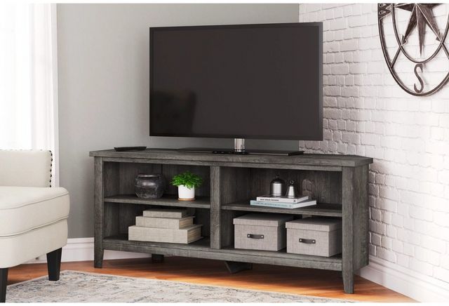 front view of a grey wood tv stand in a modern living room corner. Atop the tv stand is a tv, and in the four storage compartments are books and decor items