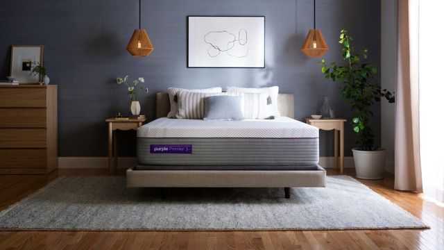 front view of a modern contemporary bedroom with a purple mattress on a beige bed frame, with pillows stacked on top. This all sits in front of a dark gray wall with white artwork.