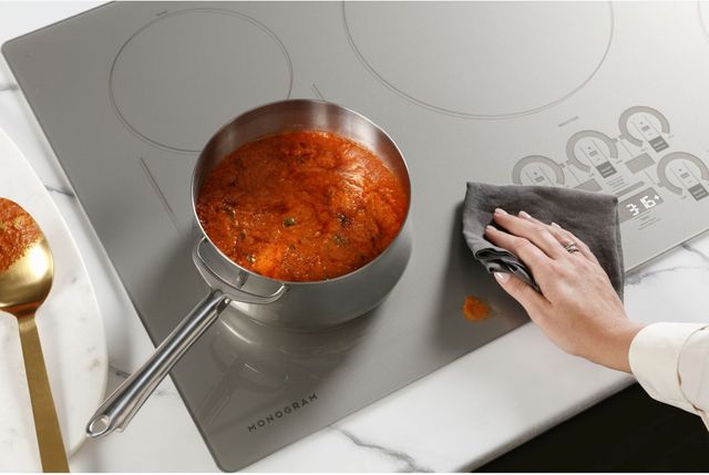 top view of a hand using a grey rag to wipe red sauce off of a monogram cooktop