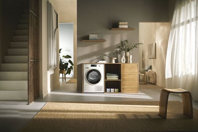 front view of a modern luxurious room with beige walls, stairs on the left, and a white heat pump dryer in a laundry console area centered in the room