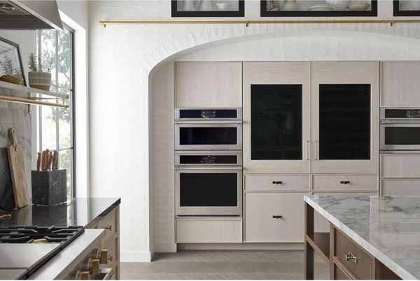 front view of a modern kitchen facing a monogram double wall oven in front, a countertop to the left, and an island to the right, all styled in white, gold, and grey colors.