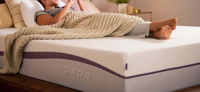side view of a woman covered in a beige knit blanket laying on a purple mattress, presumably on her cell phone which is out of shot