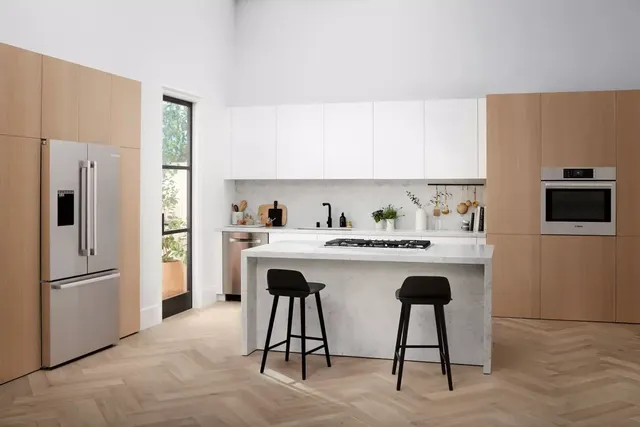 front view of a modern kitchen with light wood cabinetry and white walls, with a stainless steel counter depth refrigerator on the left