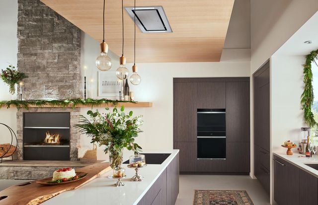 front view of a modern kitchen with an induction cooktop an island and ceiling ventilation, with a double wall oven in the background and various flowers and greenery as decor