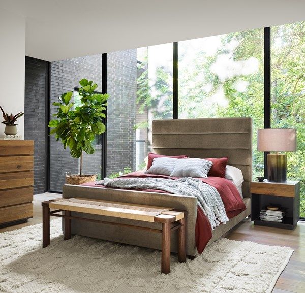 front view of a modern luxury bedroom with a beige upholstered bed frame, a wood dresser, and a small tree in a pot in the corner
