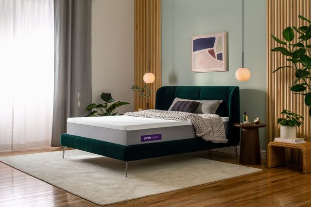 side view of a mid-century modern bedroom with an emerald green bed frame in the center. On top of the bed frame is a purple mattress with two gray pillows and a beige knit blanket. 