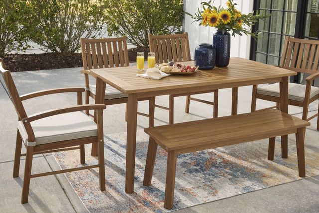 standing view of a light wood outdoor dining table with breakfast on it, and four chairs and a bench around on a stone outdoor patio 