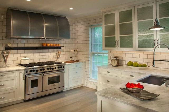 front view of a modern kitchen with white subway tile walls, white cabinets, stainless steel appliances, and a large custom range hood 